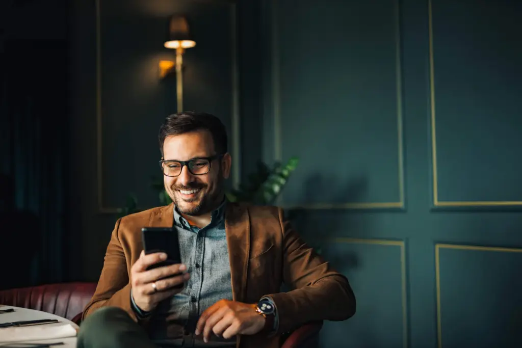 Ein Mann mit Brille und Jacket schaut lächelnd auf sein Handy in der Hand